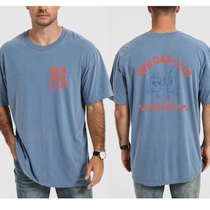 May include: A blue t-shirt with the text "NEW DAD CLUB" in orange on the front and back. The back of the shirt features a cartoon coffee cup and crayon with the text "EST. 2023" and a red and white checkered pattern.