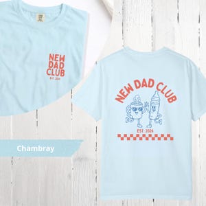 May include: Light blue t-shirts with the text "NEW DAD CLUB" in red. One shirt has the text on the front, and the other has a graphic of a coffee cup and baby bottle with the text "EST. 2026" on the back. The shirts are made of chambray fabric.