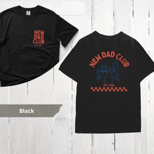 May include: Black t-shirts with the text "NEW DAD CLUB" in red. One shirt has the text on the front, the other on the back with a cartoon coffee cup and baby bottle design in blue. The shirts have the text "EST. 2026".