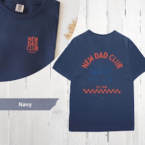May include: Navy blue t-shirt with the text "NEW DAD CLUB" in red. The shirt has a small logo on the front and a larger design on the back, including the text "EST. 2026". The design includes a red and white checkered pattern.
