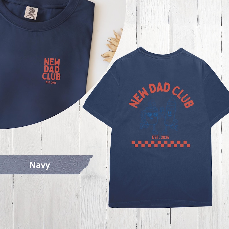 May include: Navy blue t-shirt with the text "NEW DAD CLUB" in red. The shirt has a small logo on the front and a larger design on the back, including the text "EST. 2026". The design includes a red and white checkered pattern.