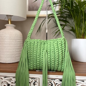 May include: A lime green crochet handbag with long fringe detailing. The bag features a rectangular body with braided handles and long fringe tassels hanging from the bottom. The bag is made of a thick, textured yarn.