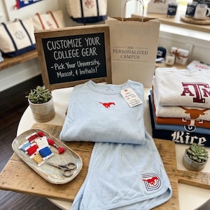 Op de afbeelding: Een display van aanpasbare college-uitrusting, waaronder lichtblauwe sweatshirts en shorts met een rood mustang-logo. De scène omvat draagtassen, wimpels en een bord met de tekst "Customize Your College Gear - Pick Your University, Mascot, & Initials!"