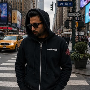 May include: A black zip-up hoodie featuring a silver zipper pull, a white Chrome Hearts logo on the left chest, and a red cross graphic on the left sleeve. The garment includes a drawstring hood, ribbed cuffs, and a front kangaroo pocket.