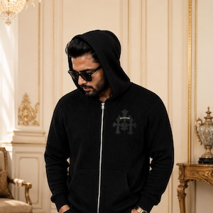 May include: A black ribbed knit zip-up hoodie featuring a hood, long sleeves, and a silver-toned metal zipper closure. The chest displays a dark, tonal cross-shaped emblem. The garment is constructed with a vertical ribbed texture throughout.