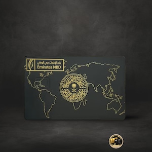 Może przedstawiać: Ciemnoszara karta kredytowa ze złotymi akcentami. Karta posiada wzór mapy świata i logo Emirates NBD. Środek karty ma ozdobny złoty emblemat.