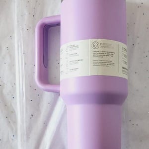 Puede incluir: Un vaso t&eacute;rmico aislado de color lavanda con asa y tapa transparente. El vaso tiene un borde de acero inoxidable y una etiqueta blanca con texto negro. Una pajita transparente es visible a la izquierda.