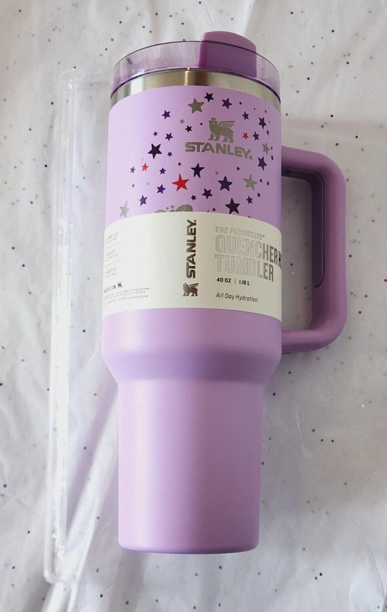 Puede incluir: Un vaso Stanley Quencher lavanda con asa, decorado con estrellas. El vaso tiene una capacidad de 1,18 L y presenta el logotipo de Stanley e informaci&oacute;n del producto.