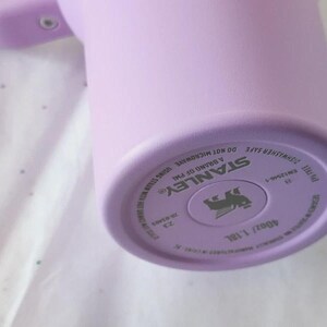 Puede incluir: Una botella de agua Stanley de color lavanda. La parte inferior de la botella muestra el logotipo de Stanley, informaci&oacute;n del producto y el texto "40oz/1.18L". La botella es de color lavanda s&oacute;lido.