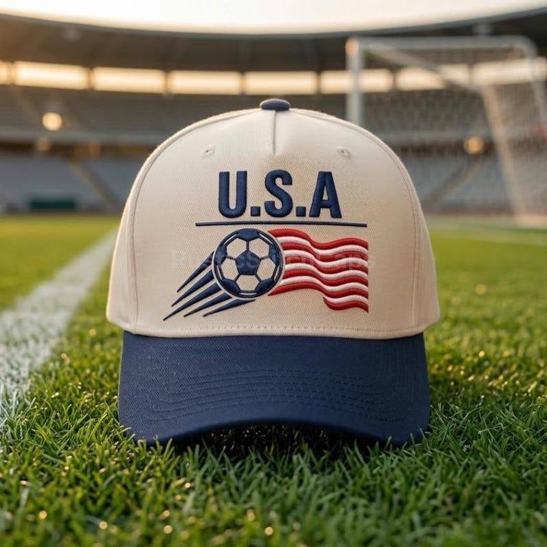 Peut inclure: Une casquette de baseball beige avec une visi&egrave;re bleu marine, orn&eacute;e d'une broderie repr&eacute;sentant un ballon de football avec des lignes de mouvement et un drapeau ondul&eacute; rouge et blanc. Le texte U.S.A est brod&eacute; en bleu marine au-dessus du motif.