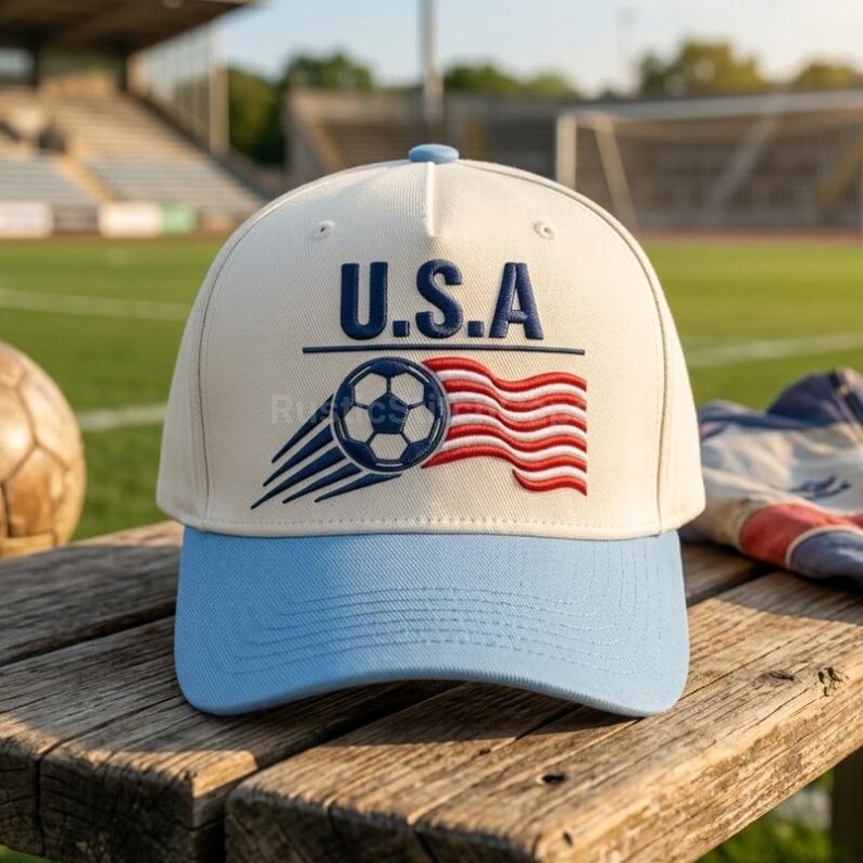 Peut inclure: Une casquette de baseball couleur cr&egrave;me avec une visi&egrave;re bleu clair. L'avant pr&eacute;sente le texte brod&eacute; U.S.A. au-dessus d'un graphique repr&eacute;sentant un ballon de football avec des lignes de mouvement et un drapeau ondul&eacute; rouge et blanc.