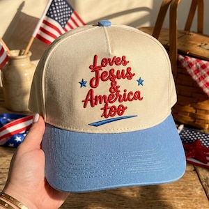 May include: A beige and light blue baseball cap with the embroidered text "Loves Jesus & America too" in red. The cap has a curved brim and a blue button on top. The background includes American flags and a picnic basket.