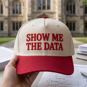 May include: A beige and red baseball cap with the text "SHOW ME THE DATA" embroidered in red. The cap has a red brim and a red button on top. The cap is held in front of a blurred background of a building and papers.