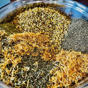 May include: A large stainless steel bowl filled with a variety of dried herbs and flowers. The bowl is divided into sections of yellow, green, and grey plant matter, including chamomile flowers and other dried leaves.