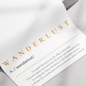 Peut inclure: Une feuille de papier blanche avec le mot "WANDERLUST" imprimé en or. Le papier contient une définition en dessous. Il est légèrement courbé et posé sur une surface blanche, avec des ombres.