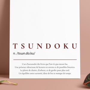 Peut inclure: Une affiche blanche avec le mot "TSUNDOKU" en texte bordeaux. En dessous, la définition et une courte description en français. Deux feuilles aux motifs verts et oranges sur fond rose.