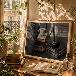 Peut inclure: Une impression encadrée en noir et blanc avec le mot "Tsundoku" et sa définition en français. L'impression est posée sur un bureau en bois avec des livres, une tasse, un appareil photo et une bougie allumée. Des plantes vertes et une fenêtre avec vue sur la ville sont en arrière-plan.