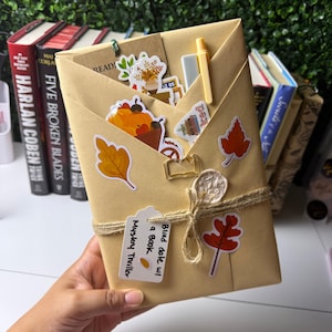 Puede incluir: Un libro envuelto en papel color canela con un cordel, un sello de cera y pegatinas otoñales de hojas y calabazas. Una etiqueta dice "Blind date w/ a book Mystery Thriller". Incluye un clip dorado con forma de libro y un pequeño bolígrafo.