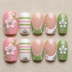 May include: A set of artificial nails in various designs. Pink nails with white flowers and rhinestones, white nails with pink and green stripes, and nails with a pink base, green accents, and rhinestones. Green nails with white flowers are also included.