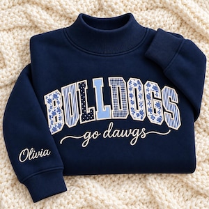May include: A navy blue mock neck sweatshirt featuring the word BULLDOGS in a mix of blue and white patterned letters, with the text go dawgs in white script below. The name Olivia is printed in white on the left sleeve cuff.