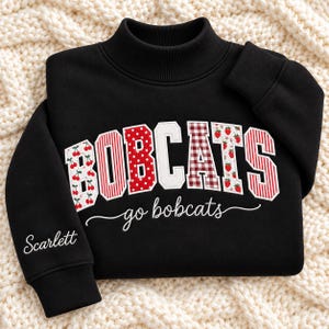 May include: A black long-sleeve sweatshirt with a mock neck. The front features the word BOBCATS in large, patterned block letters with cherry, polka dot, and strawberry prints. Below, the text go bobcats is written in white script. The name Scarlett is embroidered on the sleeve.