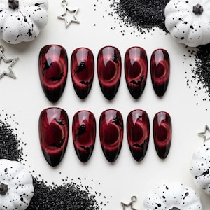 May include: A set of ten oval-shaped press-on nails. Each nail features a dark red base with a swirling, lighter red design, and black silhouettes of bats and ghosts. The nails are arranged on a white surface with Halloween-themed decorations.