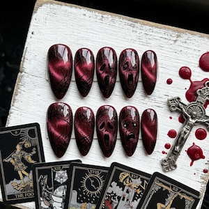 May include: A set of ten burgundy-colored, oval-shaped press-on nails. Some nails feature a textured design, while others display a screaming face graphic. Tarot cards and a crucifix are also present.