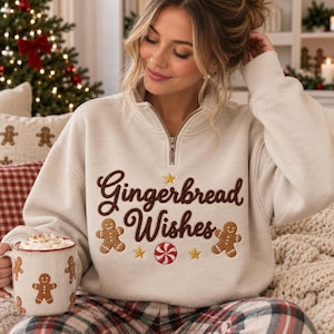 May include: A beige quarter-zip sweatshirt featuring embroidered brown text that reads Gingerbread Wishes, accompanied by two gingerbread man figures, three yellow stars, and a red and white peppermint candy graphic on the front.