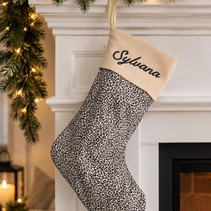 Könnte beinhalten: Ein Weihnachtsstrumpf mit einem schwarz-weißen Leopardenmuster auf dem Hauptteil und einem beigen Umschlag am oberen Rand. Der Name Sylvana ist in schwarzer Schreibschrift auf den Umschlag gestickt, und eine goldene Bandschlaufe ist befestigt.