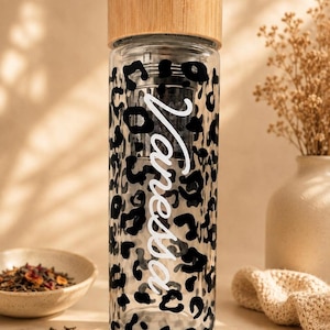 Könnte beinhalten: Eine klare Glasflasche mit schwarzem Leopardenmuster und dem Namen Vanessa in weißer Schreibschrift. Die Flasche enthält ein zylindrisches Teesieb aus Metall und ist mit einem hellen Schraubverschluss aus Holz versehen.