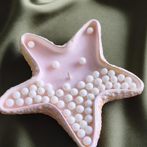May include: A light pink, starfish-shaped candle featuring a textured outer edge and a smooth interior surface. The candle is decorated with numerous small, round, white pearl-like beads embedded across the surface, with a single central wick.
