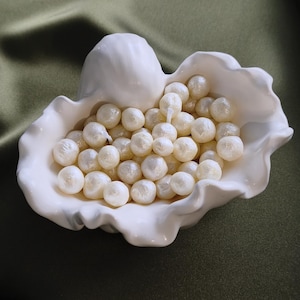 Peut inclure: Un bol en céramique blanche en forme de coquillage rempli de nombreuses petites bougies rondes couleur crème ressemblant à des perles. Chaque bougie présente un léger éclat et une mèche visible, formant un amas de sphères de cire nichées dans les bords incurvés du récipient blanc brillant.