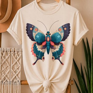 May include: Cream-colored t-shirt featuring a large butterfly design. The butterfly has blue, teal, pink, and orange details. The shirt is knotted at the waist and hangs on a wooden hanger.