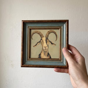 Könnte beinhalten: Ein quadratisch gerahmtes Gemälde einer Ziege mit geschwungenen Hörnern, beigem und schwarzem Fell sowie nach vorne gerichteten Augen. Das Kunstwerk befindet sich in einem dekorativen Rahmen mit blau-grauem Innenrand und goldener Perlenleiste.