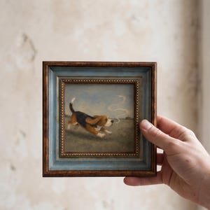 Puede incluir: Un cuadro cuadrado enmarcado que muestra a un perro beagle marrón, negro y blanco corriendo por un campo. El perro aparece en movimiento con una estela de humo blanco arremolinado detrás. El marco es de madera oscura con borde azul y cuentas doradas.