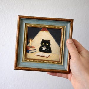 以下が含まれることがあります： 額縁に入ったアートワーク。黒猫が机でマグカップから飲み物を飲んでいます。猫は暖かい光の下にいて、近くには本とろうそくが灯っています。フレームは青と金の縁取りです。