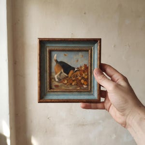 Peut inclure: Une petite peinture carrée encadrée représentant un chien beagle au pelage noir, blanc et fauve reniflant un tas de feuilles d'automne orange et brunes. Le cadre présente un bord extérieur en bois foncé et une bordure intérieure bleu clair dorée.