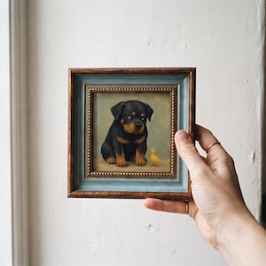 May include: A square framed oil painting featuring a black and tan Rottweiler puppy sitting next to a small yellow chick. The frame is constructed with a dark brown outer edge and a light blue inner border, accented by a gold beaded trim.