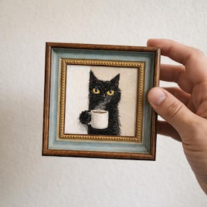 以下が含まれることがあります： 黄色い目を持ち、白いコーヒーマグを持つ黒猫の額入り絵画。 フレームは茶色と青色です。 このアートワークはユーモラスな品質を持ち、少し不機嫌そうな表情の猫を描いています。