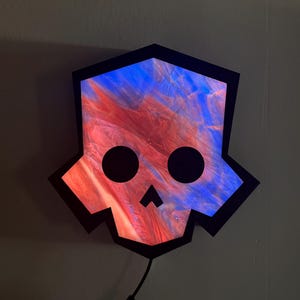 May include: A skull-shaped wall art piece with a black outline and a colorful, abstract design in red and blue. The eyes are represented by two large black circles, and the mouth is a small black triangle.