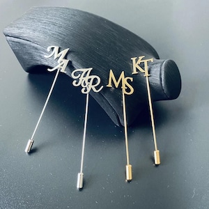 Op de afbeelding: Vier decoratieve reversspelden met de initialen "M A", "Mr", "M S" en "K T". De spelden zijn in zilver- en goudtinten, met een lange speld aan de achterkant. De spelden worden gepresenteerd op een zwart oppervlak.