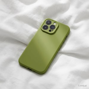 Puede incluir: Una funda de smartphone de color verde oliva mate con un módulo de cámara cuadrado elevado que presenta tres lentes circulares y un pequeño flash. La funda tiene un acabado liso de color sólido y lleva la palabra Umniyat en la esquina inferior.