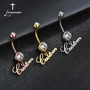 Op de afbeelding: Drie navelpiercings in roségoud, goud en zilver. Elke ring heeft een heldere edelsteen en een bungelend "Custom"-script. De merknaam "Jetdiffery" staat linksboven.