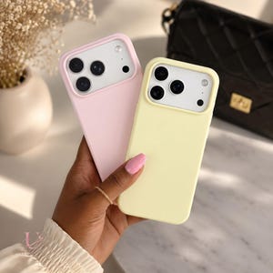 May include: Two rectangular smartphone cases, one in a soft pastel pink and the other in a pale yellow. Each case features a raised camera cutout area with three circular lens openings and a small flash hole, designed for a modern mobile device.