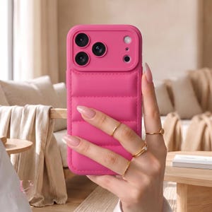 May include: A bright pink smartphone case with a quilted, puffer-style texture on the back. The case features a raised camera module cutout with three circular lens openings and a flash cutout, all encased in a matching pink frame.