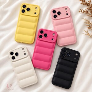 May include: Five smartphone cases with a quilted, puffer-style texture. The cases are available in yellow, light pink, hot pink, off-white, and black. Each case features a raised camera cutout section and a smooth, padded back design.