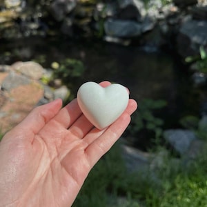 May include: A smooth, heart-shaped object with a pearlescent white finish. The surface has a subtle, shimmering quality that reflects light, giving it a soft, glowing appearance. The heart is rounded and solid, featuring a clean, polished texture.