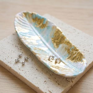 May include: A decorative, feather-shaped ceramic dish with a gold leaf design and a light blue and white background. Two pairs of silver earrings rest on the dish and a stone surface.