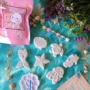May include: A plaster painting kit featuring sea-themed shapes like a mermaid tail, starfish, and seahorse. The kit includes a paint palette, paintbrushes, and a pink bag with a cartoon bunny and bear design. The background is a blue and turquoise surface.