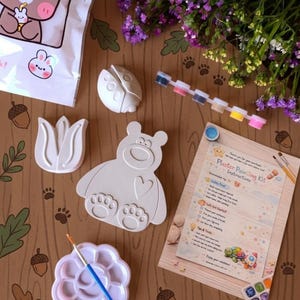 Könnte beinhalten: Ein Gipsmalset mit bären-, marienkäfer- und tulpenförmigen Gipsfiguren. Das Set enthält eine Farbpalette, Farbtöpfe und Anweisungen. Der Hintergrund ist eine Holzoberfläche mit Pfotenabdrücken und floralen Akzenten.